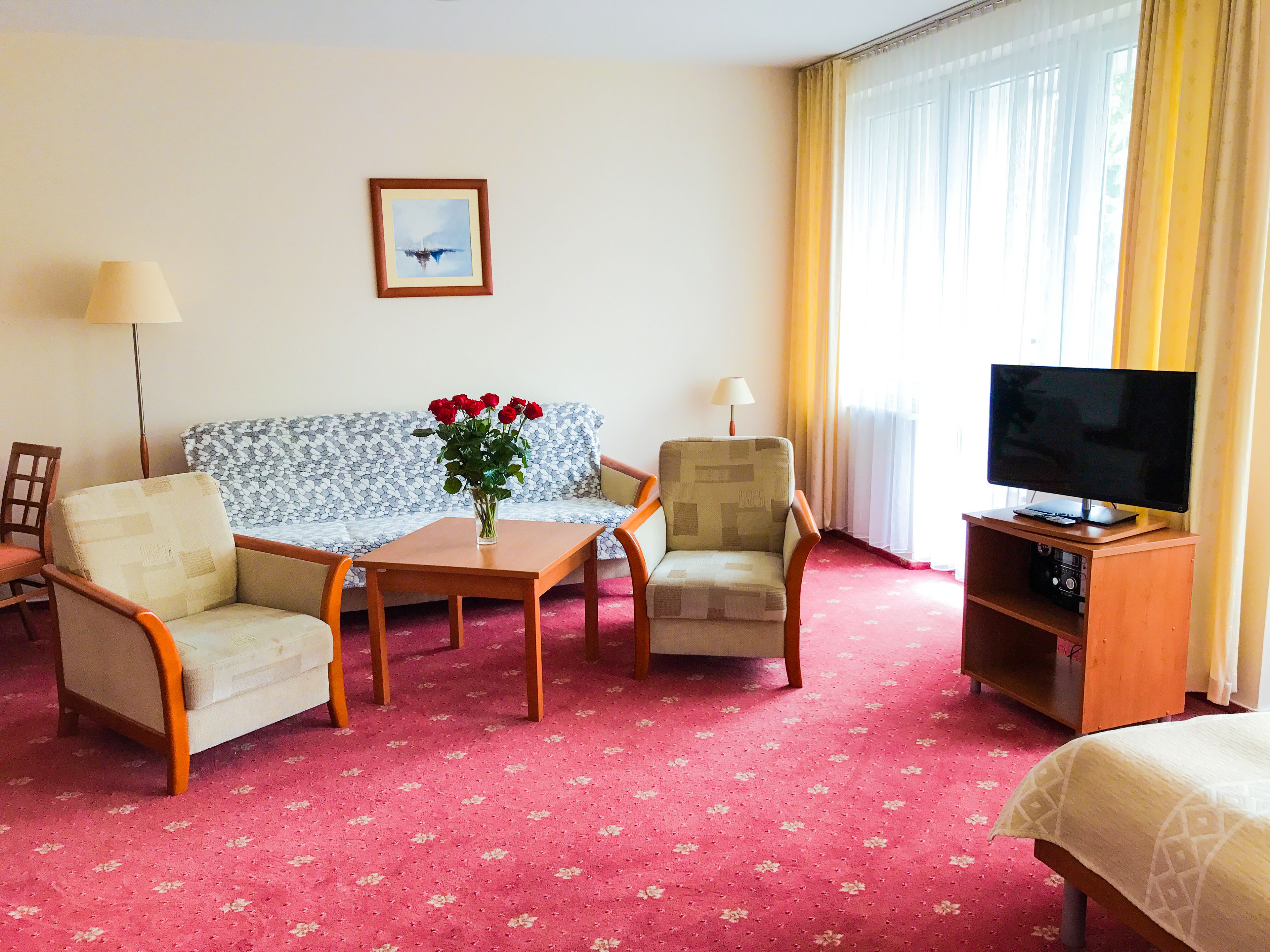 A hotel room with a sofa, two armchairs, a small table with a vase of flowers, a TV on a wooden stand, and a carpeted floor.