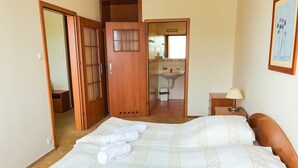Une chambre d’hôtel avec un lit, une table de chevet avec une lampe, une salle de bain visible à travers une porte ouverte et un tableau encadré au mur.