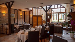 Restaurante com paredes de tijolo, vigas de madeira e janelas amplas. Mesas com toalhas brancas e cadeiras.