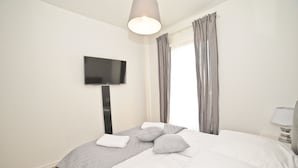 A neatly made bed with pillows and a blanket, a flat-screen TV mounted on the wall, and a window with curtains. - Grand Apartments - Modern Old Town (Gdansk)