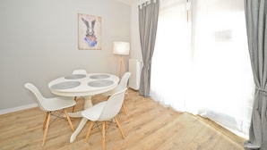 A dining area with a round table, white chairs, a painting on the wall, and a window with curtains. - Grand Apartments - Modern Old Town (Gdansk)