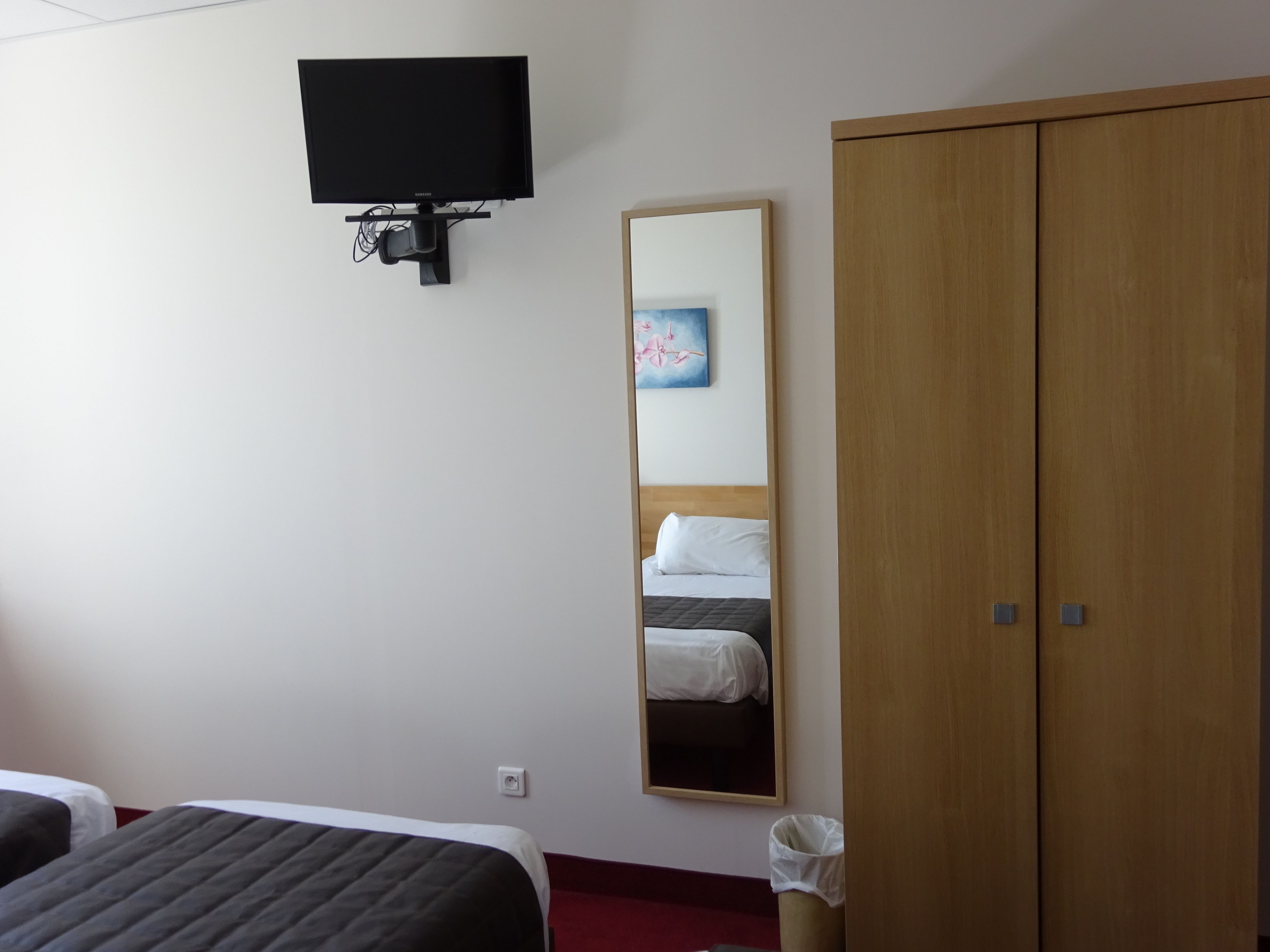 Habitación de hotel con cama, armario, televisor montado en la pared y espejo que refleja una cama y un cuadro.