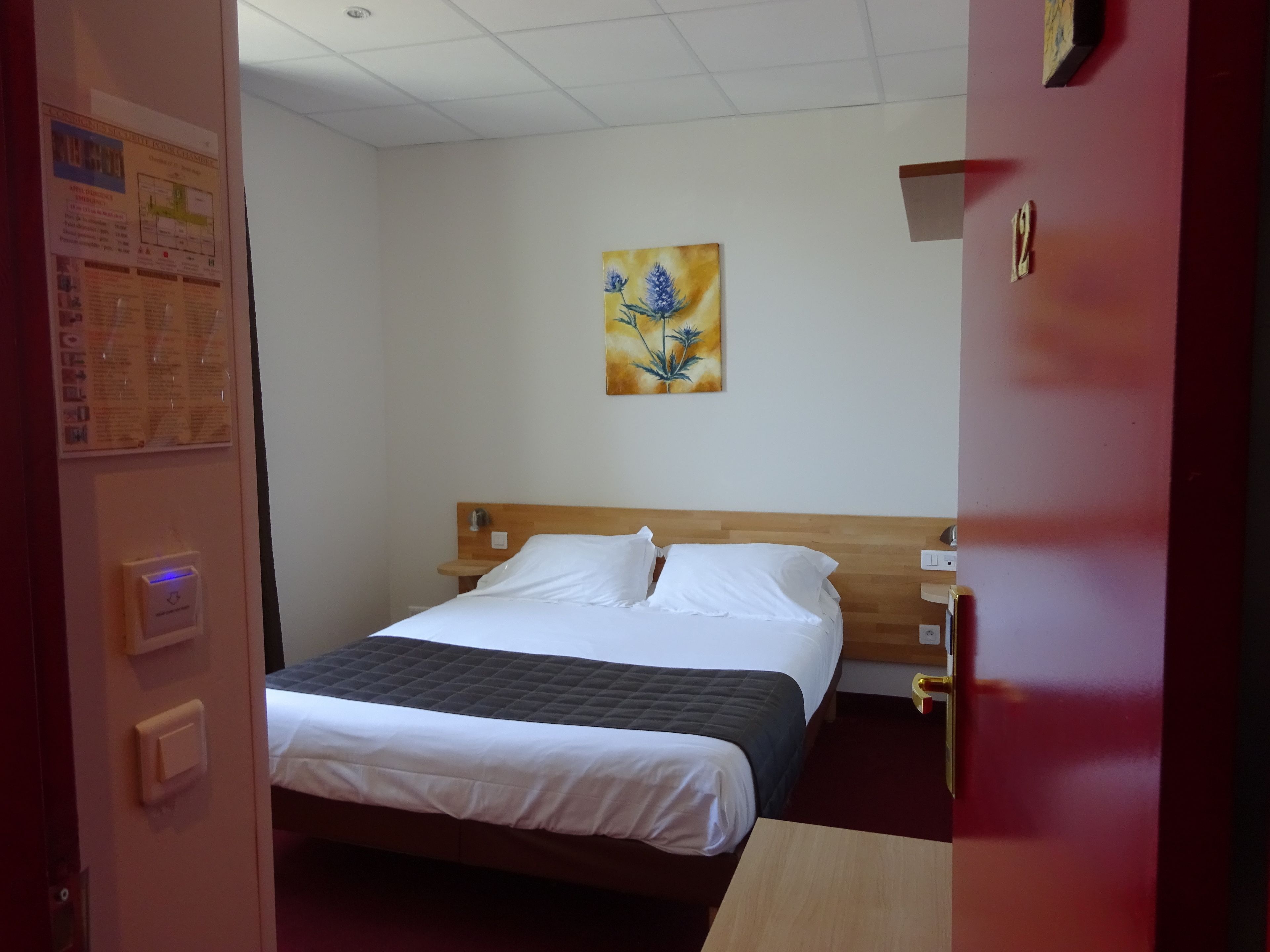 Quarto de hotel com cama, mesa de cabeceira, pintura pendurada na parede e porta com número.