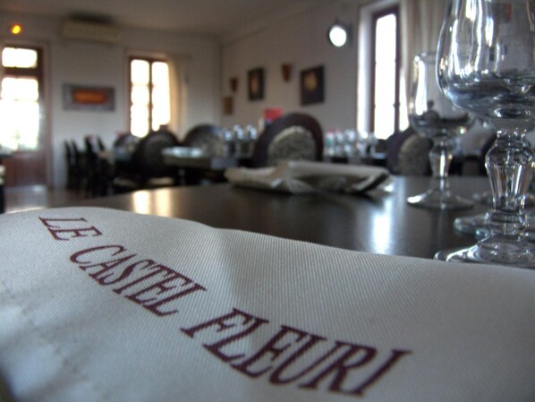 Un interior de restaurante con mesas preparadas para comer, un menú con la leyenda 'LE CASTEL PLEURI' y copas de vino sobre la mesa.