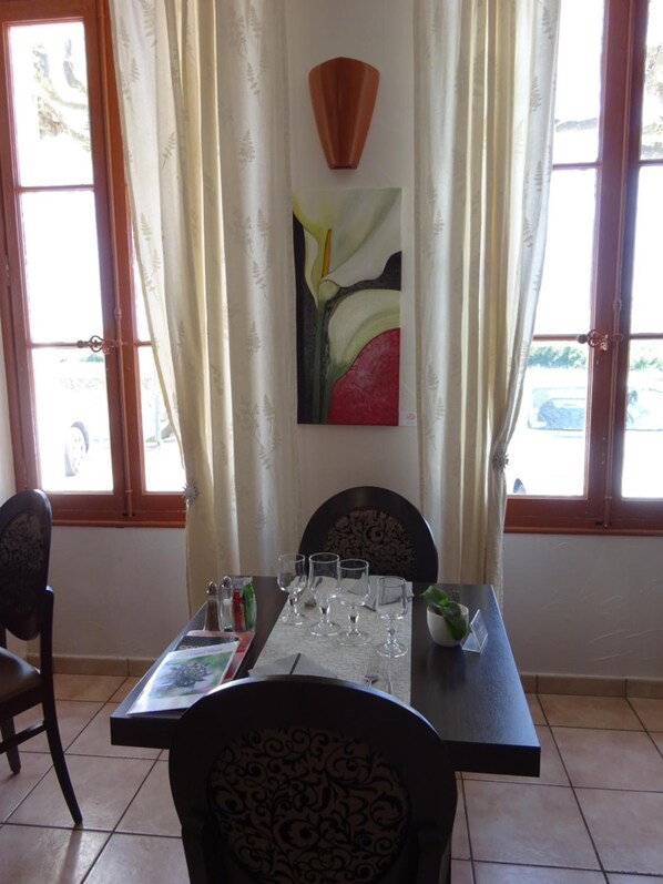 A dining area with a table set for two, a painting above the table, and a view of the street through the windows. - Hôtel Restaurant Le Castel Fleuri (Carnoules)