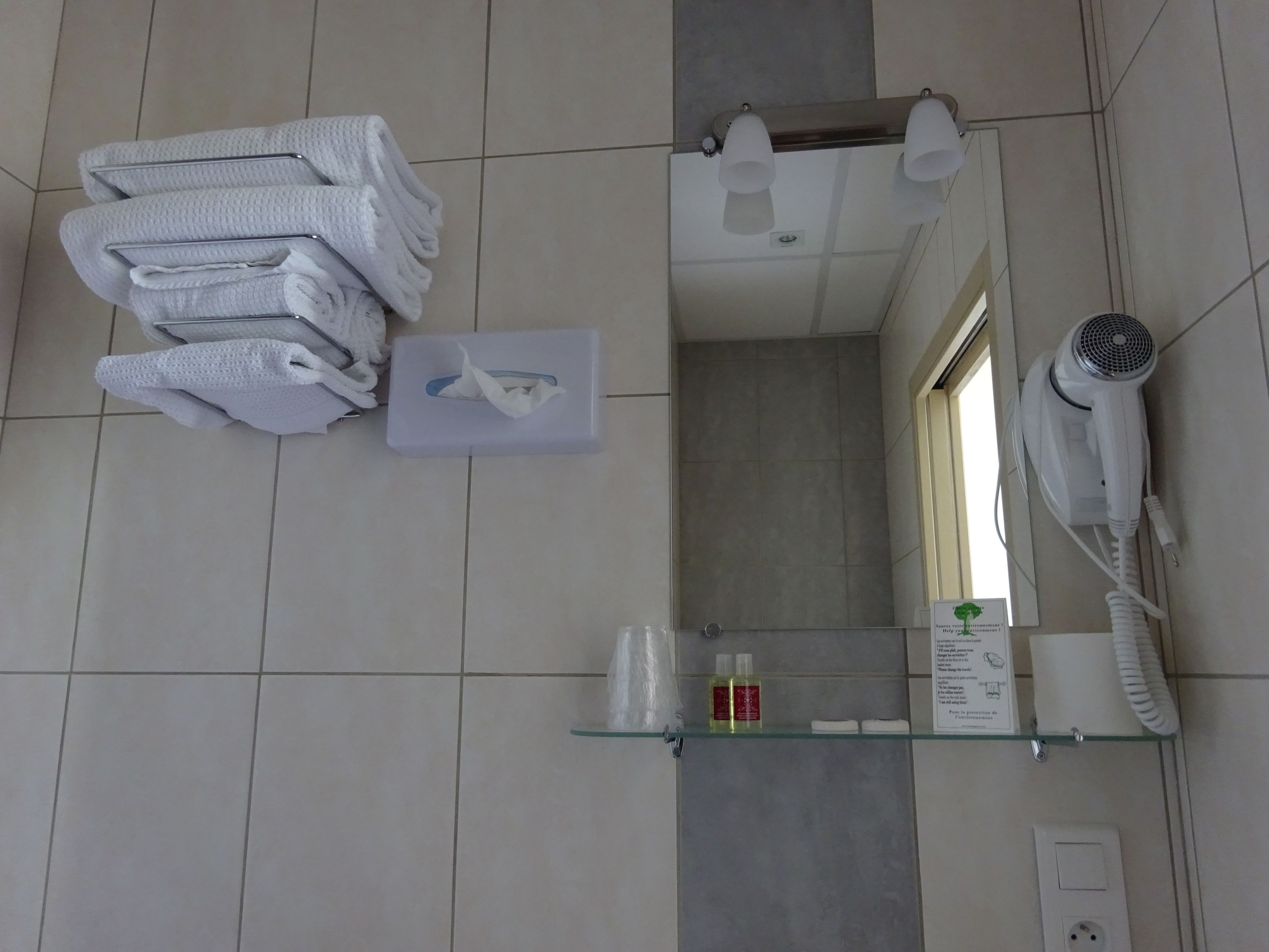 A bathroom with a stack of towels, a hand dryer, a glass shelf with toiletries, and a mirror.