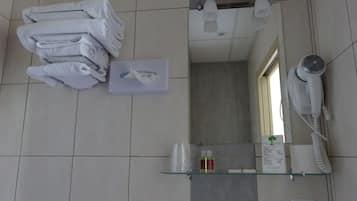 A bathroom with a stack of towels, a hand dryer, a glass shelf with toiletries, and a mirror.