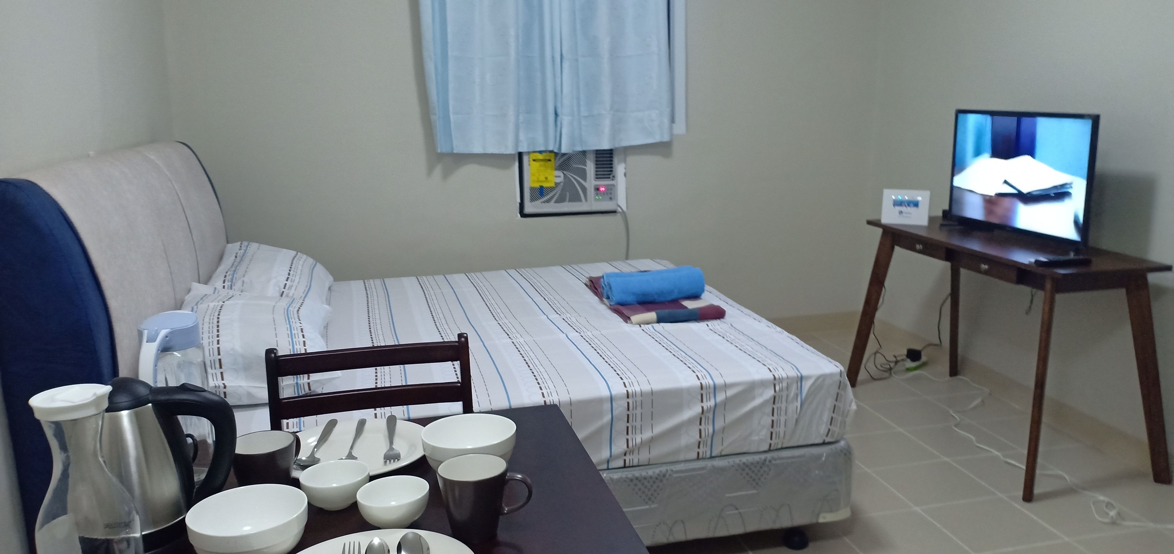 A neatly arranged room with a bed, a small table with dining items, a television, and a window with curtains.