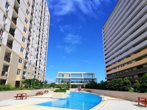 Un hotel moderno con zona de piscina, rodeado de vegetación y un cielo azul brillante.