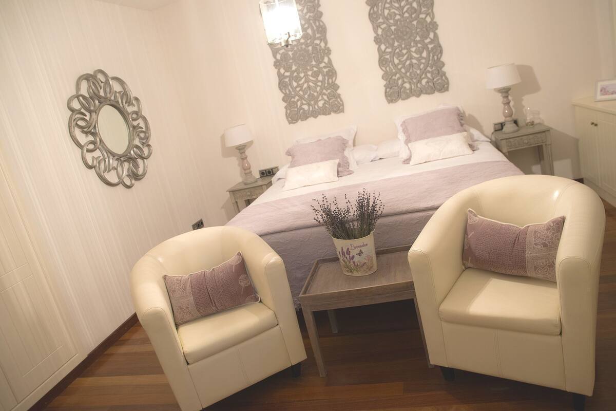 Un dormitorio con cama, dos sillas, una mesita de noche, una lámpara y decoración mural.