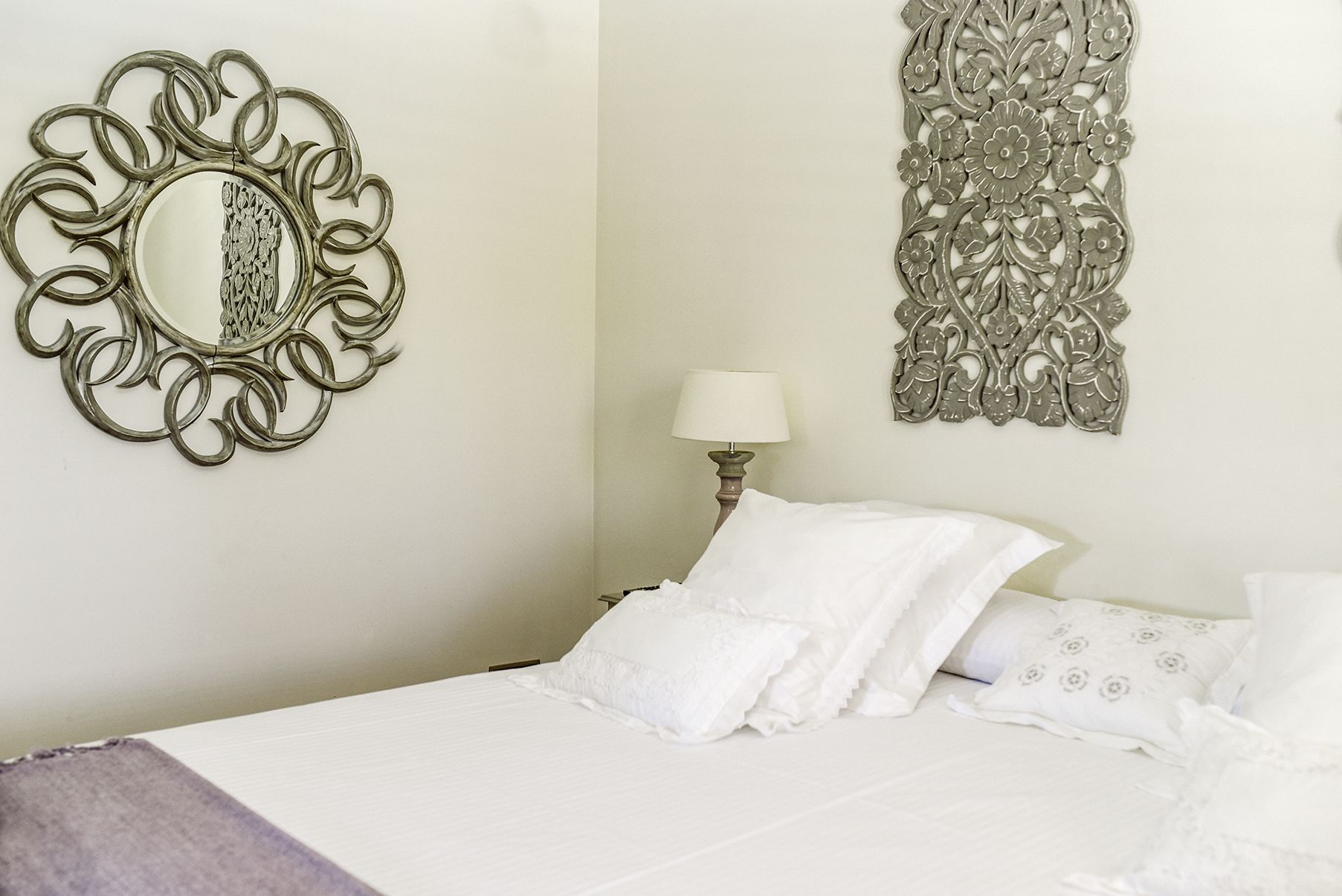 A neatly made bed with white linens and pillows, a wall-mounted mirror with an ornate frame, and a decorative wall hanging with floral patterns.