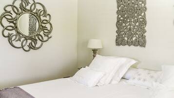 A neatly made bed with white linens and pillows, a wall-mounted mirror with an ornate frame, and a decorative wall hanging with floral patterns.