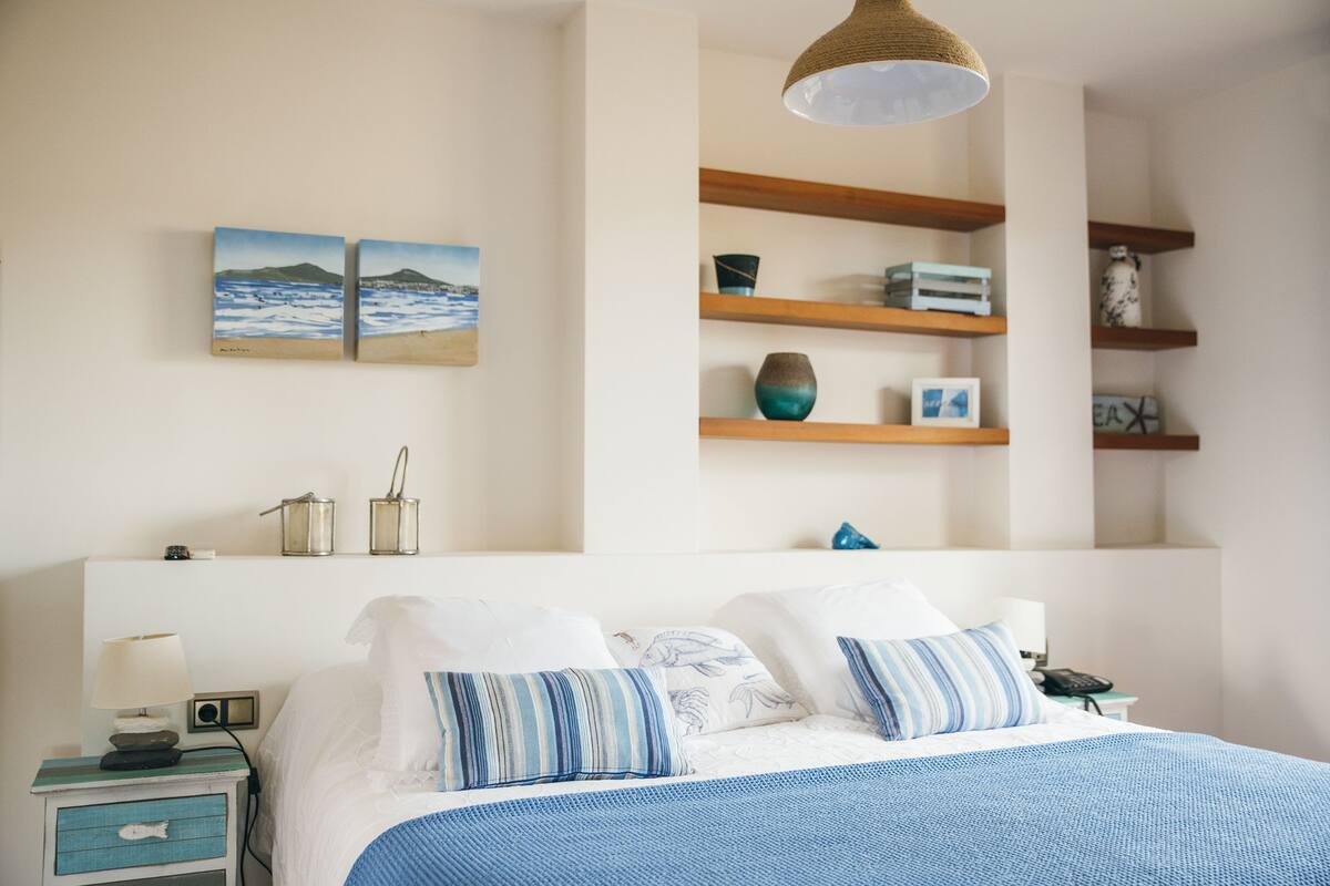 Un dormitorio ordenado con cama, mesitas de noche, lámpara y estanterías con adornos.