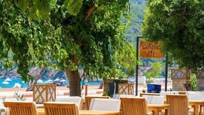 Mesas y sillas de madera al aire libre dispuestas en una terraza con vista a una piscina y una playa al fondo.