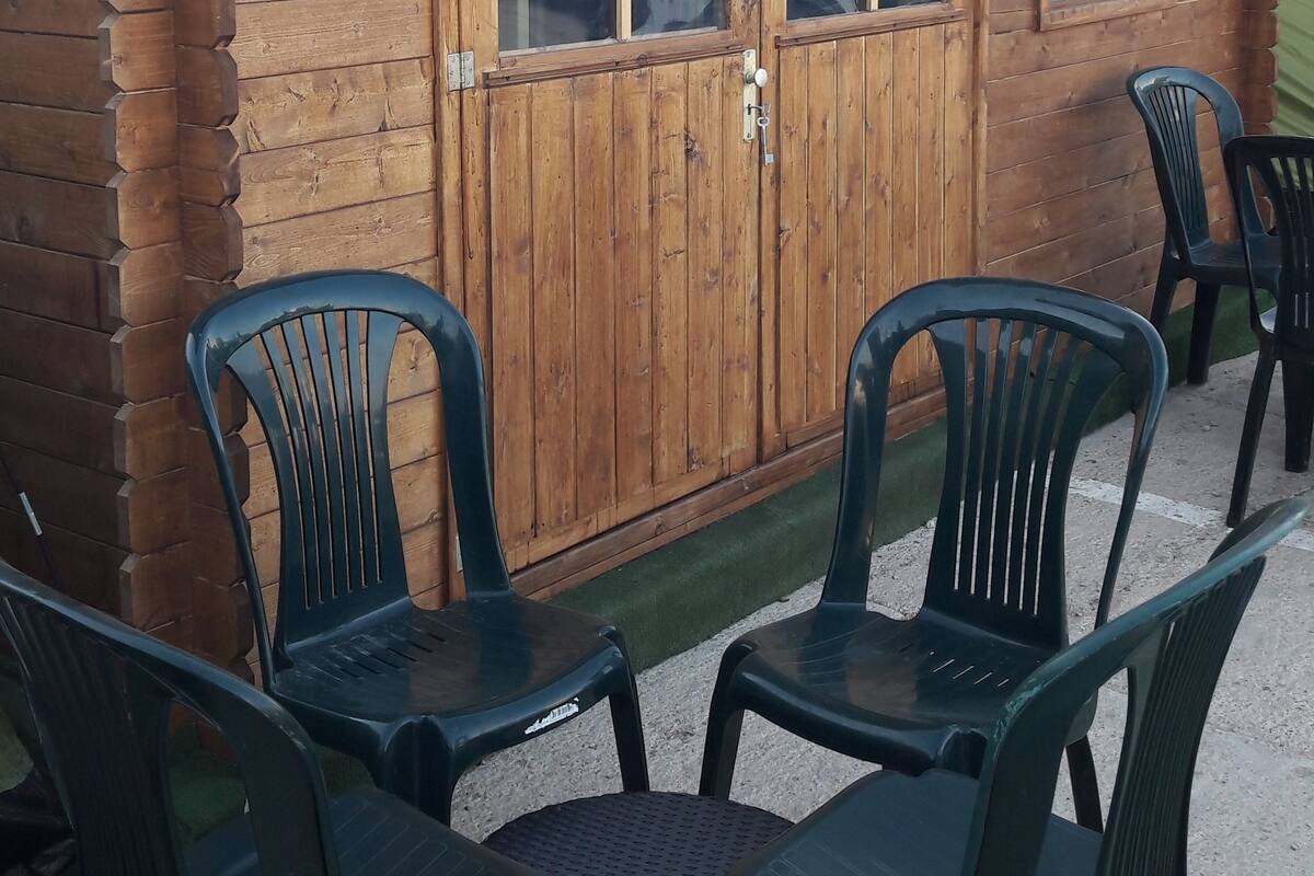 A wooden cabin with a number '1' sign, a door, and two windows. There are four green plastic chairs and a round table in front.