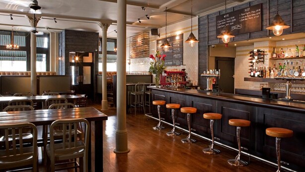 A bar area with wooden tables and stools, a bar counter with a wine rack, and a menu board above the bar. - The Tommyfield (London)