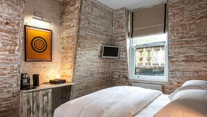 A room with a rustic brick wall, a bed with white bedding, a mounted TV, and a window with a view of outdoor statues. - The Tommyfield (London)