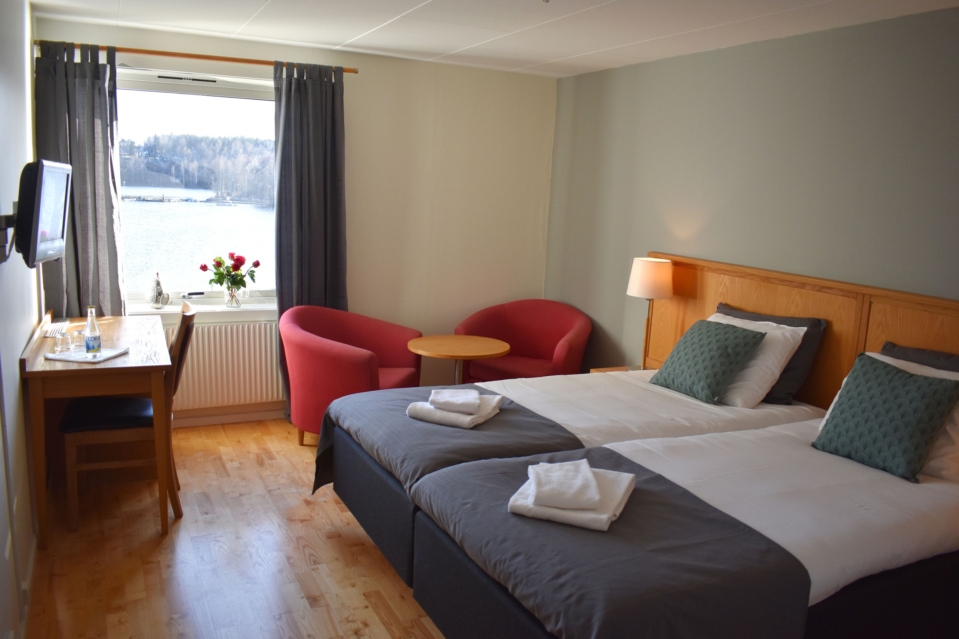 A hotel room with two beds, a desk with a chair, a small table, and a view of a body of water through a window.