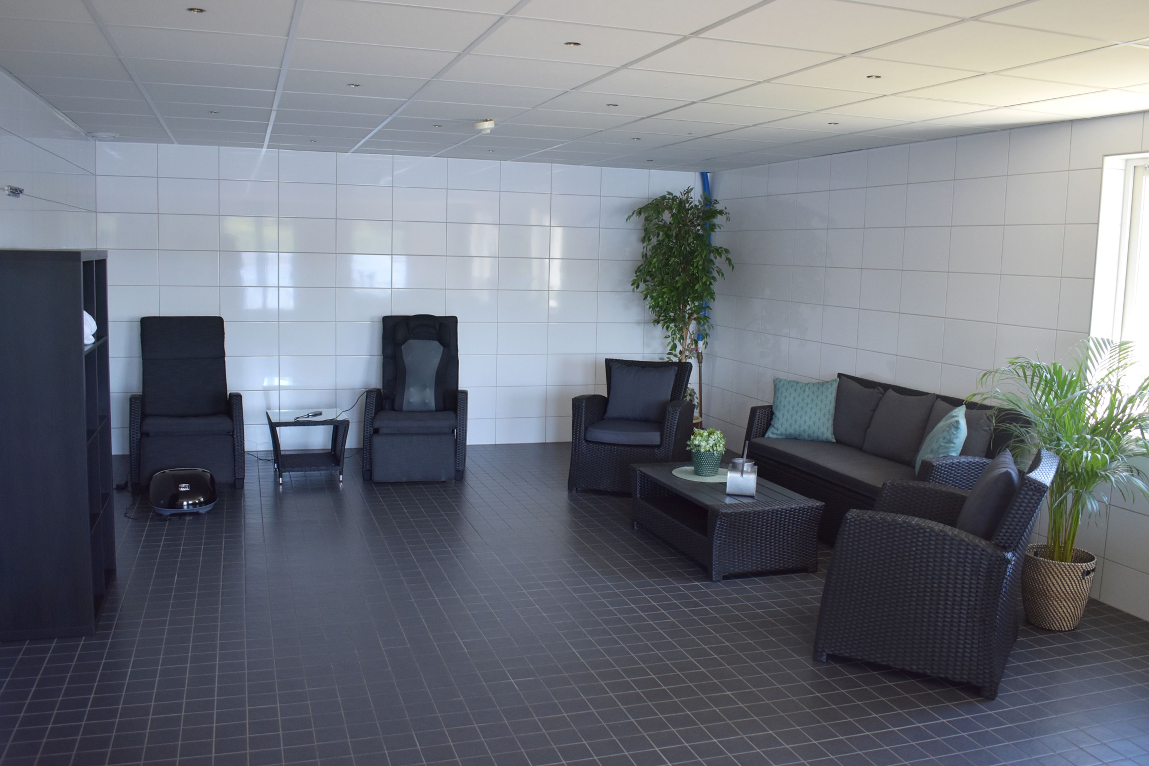A spacious room with a dark floor, white tiled walls, and a large window. There are several dark-colored chairs and a sofa arranged for seating.