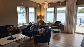 Een moderne woonkamer met uitzicht op een meer, met blauwe fauteuils, een rond houten poef en een kroonluchter.