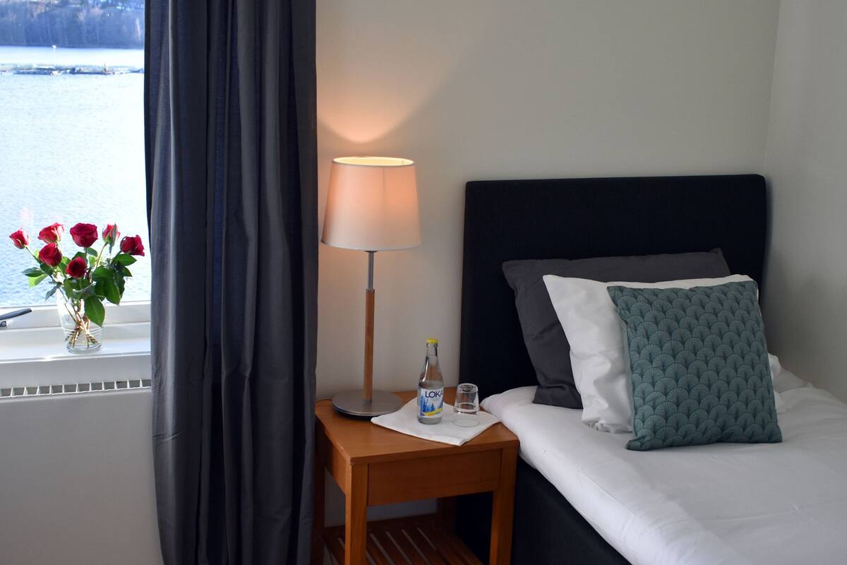 Quarto de hotel com uma cama, mesa de cabeceira com abajur, vaso com flores e vista para a água lá fora.