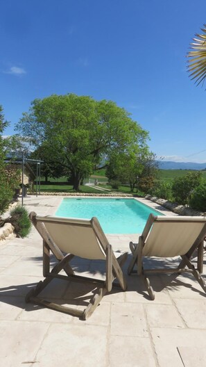 Una piscina con dos sillones de madera frente a ella, rodeada por un patio de piedra y abundante vegetación.