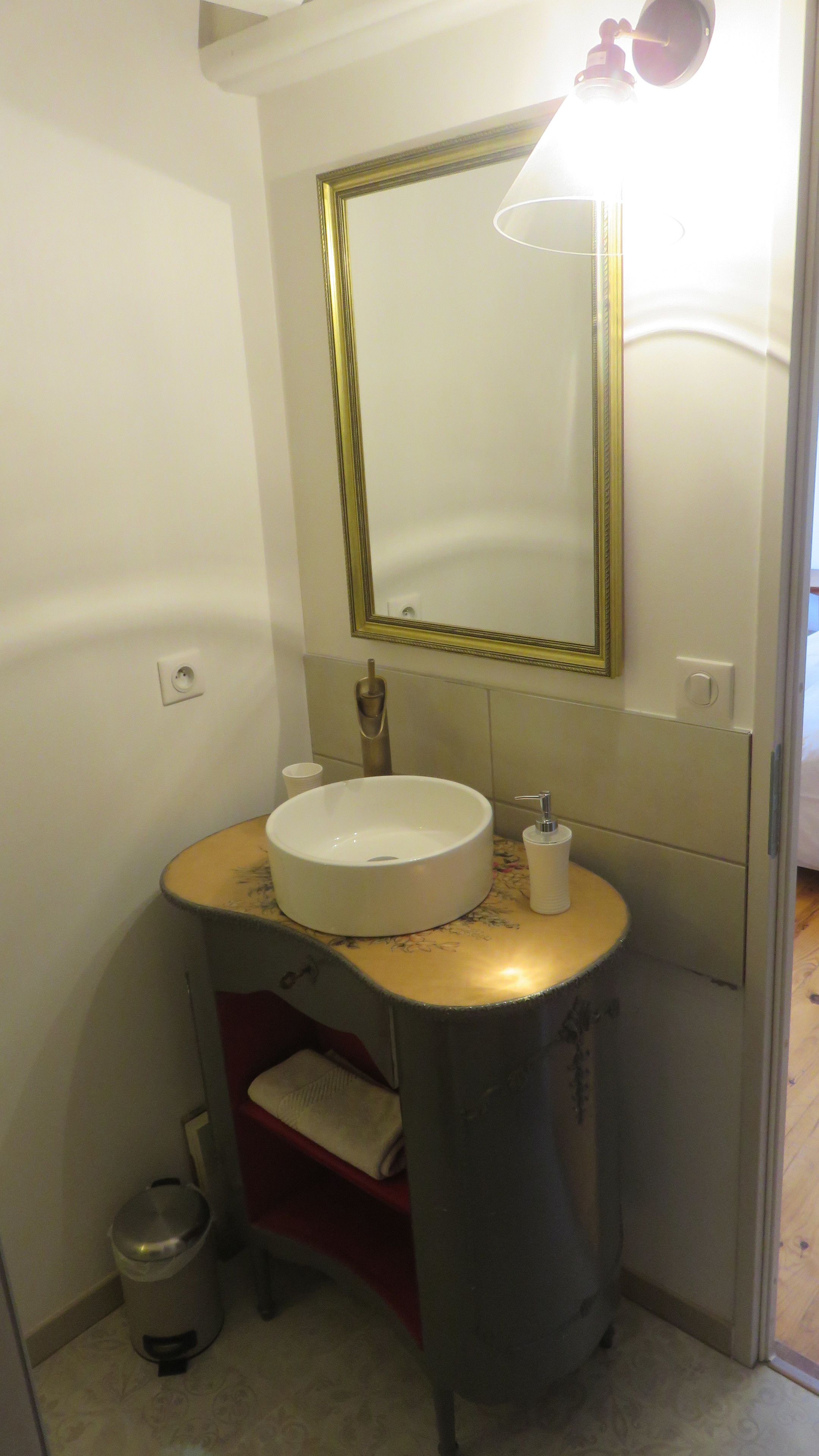 A bathroom with a white sink, a mirror with a gold frame, a towel rack, and a trash can.