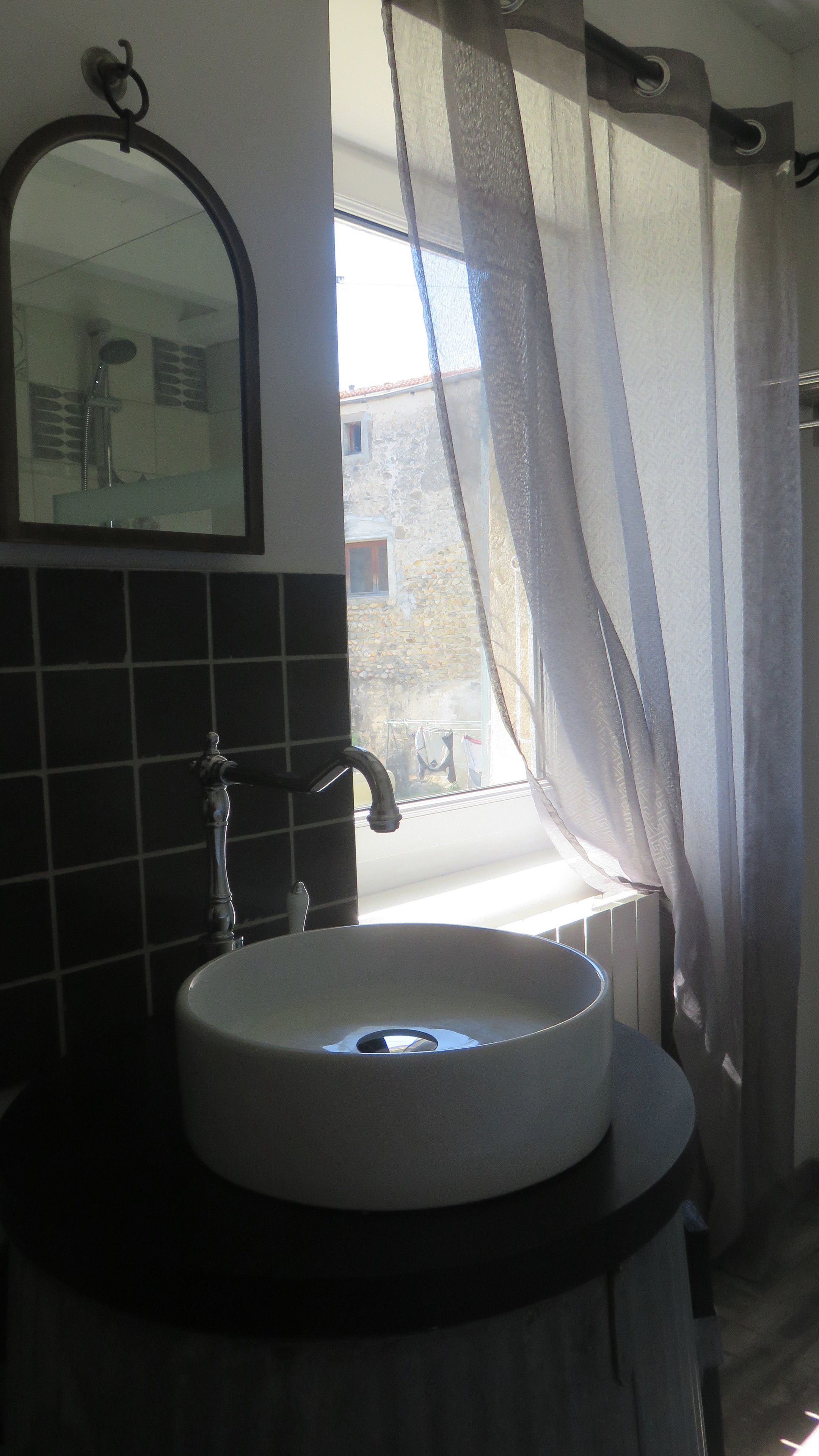 Un baño con un lavamanos redondo, un espejo y una ventana con cortinas de encaje.