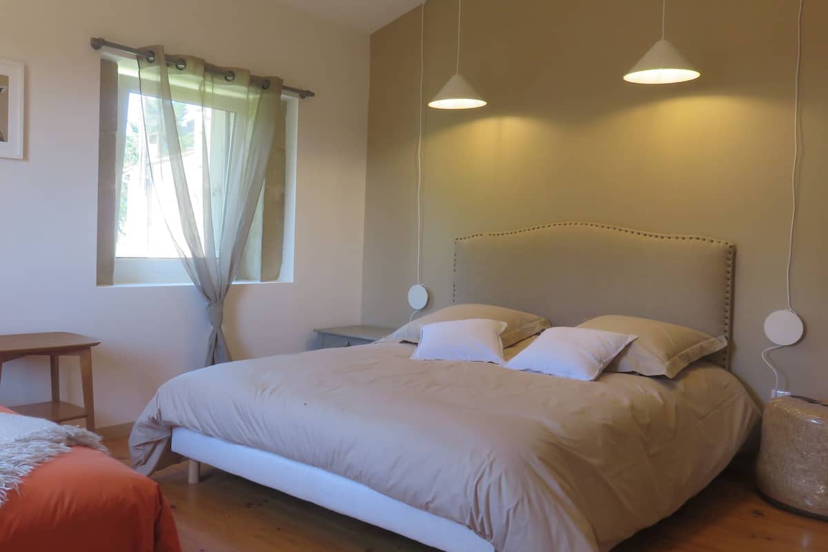 A bedroom with a bed, bedside tables, a chair, a window with curtains, and a wall-mounted lamp.
