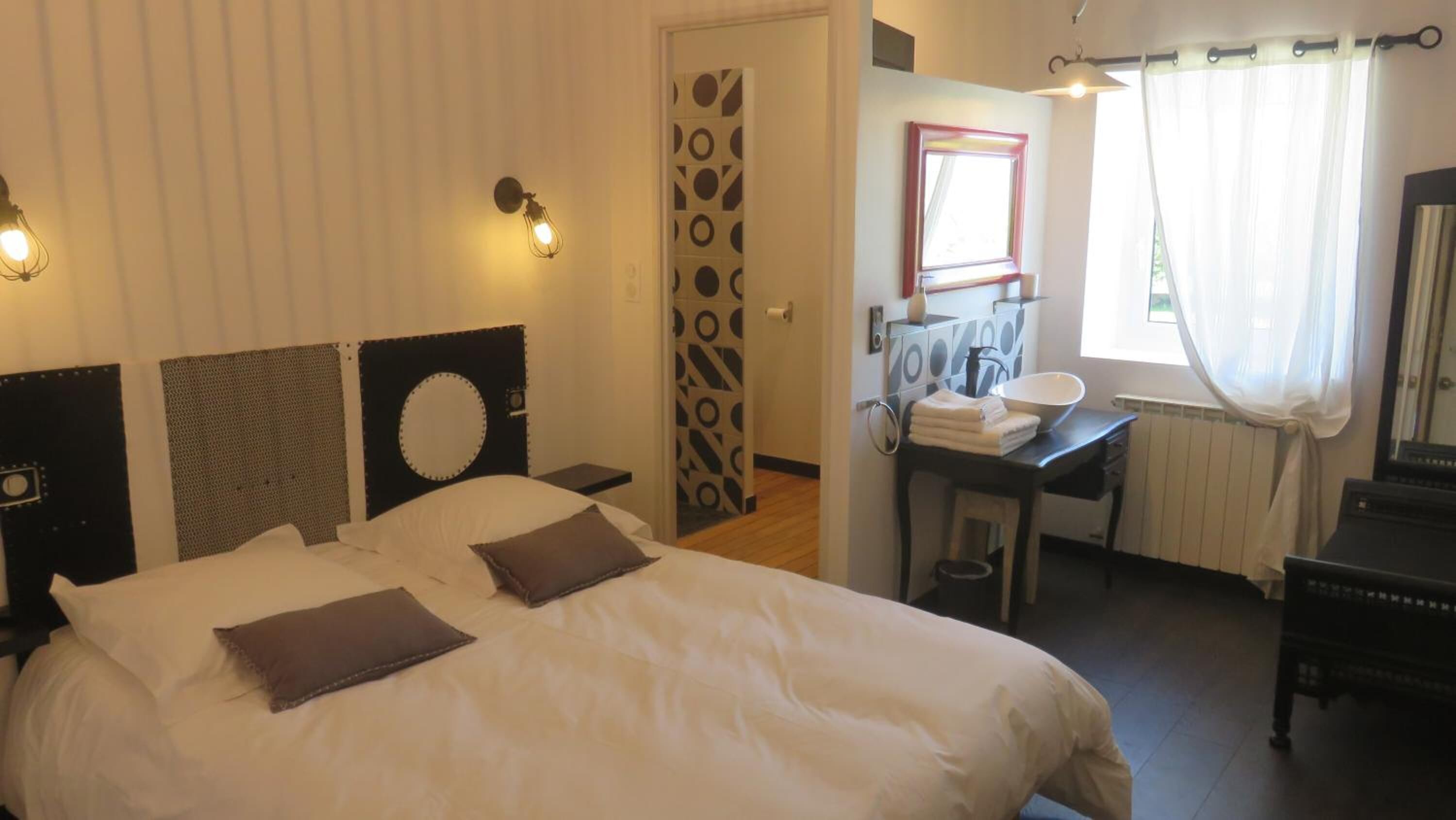 A bedroom with a bed, bedside tables, a mirror, a window with curtains, and a bathroom with a sink and towel rack.