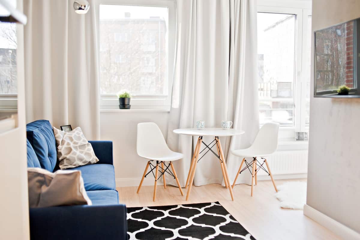 Ein modernes Wohnzimmer mit einem blauen Sofa, zwei weißen Sesseln, einem kleinen Tisch und einem Fenster mit Vorhängen.
