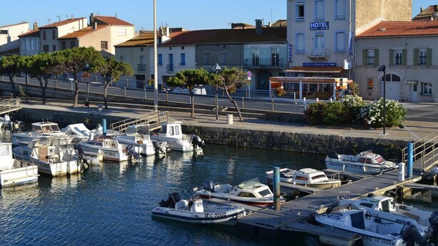 A marina with several boats docked, a hotel building with a sign, and a street with buildings and trees. - L'Hôtel du Port (Port-la-Nouvelle)