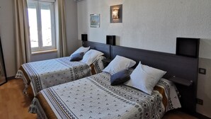 A hotel room with two beds, a window with curtains, an air conditioning unit, and a wall-mounted light fixture. - L'Hôtel du Port (Port-la-Nouvelle)