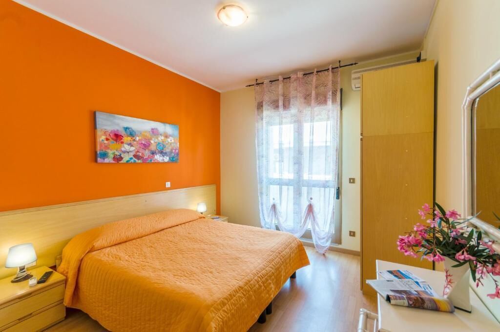 A bedroom with an orange wall, a bed with a matching blanket, a nightstand with a lamp, a painting, a window with sheer curtains, and a mirror with a vase of flowers.