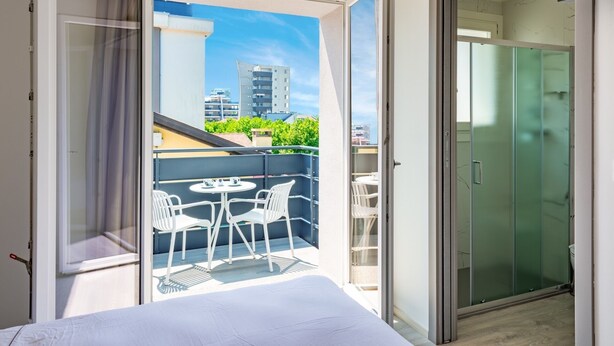 Habitación de hotel con balcón, una cama, una mesa y sillas, y una puerta corrediza de vidrio.