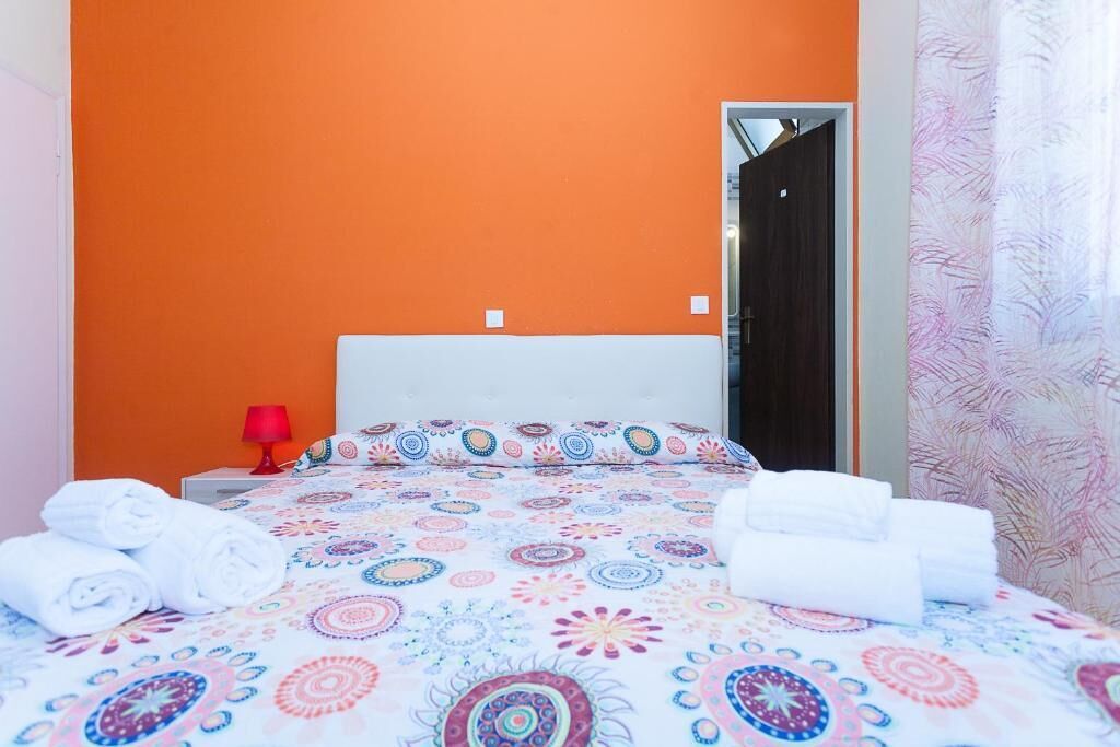 A hotel room with an orange accent wall, a bed with a colorful patterned bedspread, a red lamp, and neatly folded towels.