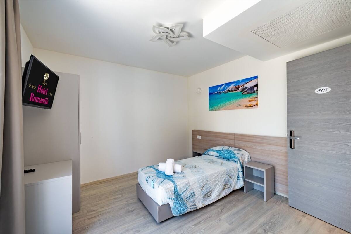 Une chambre d’hôtel avec un lit, une table de chevet, un téléviseur fixé au mur et une porte portant un numéro.