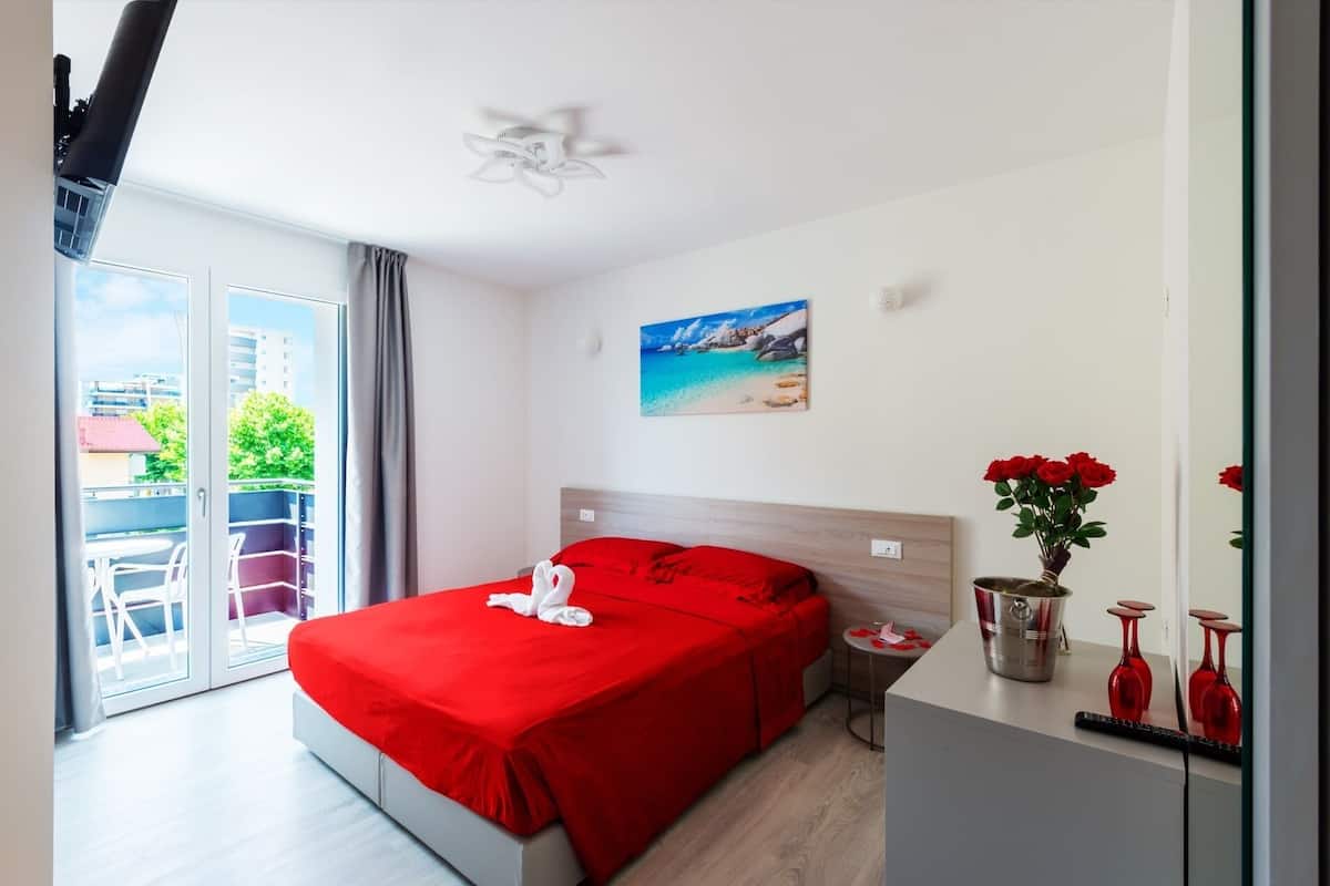 Une chambre d’hôtel avec un grand lit, un balcon avec des chaises, un tableau représentant une plage et un vase rempli de fleurs rouges.