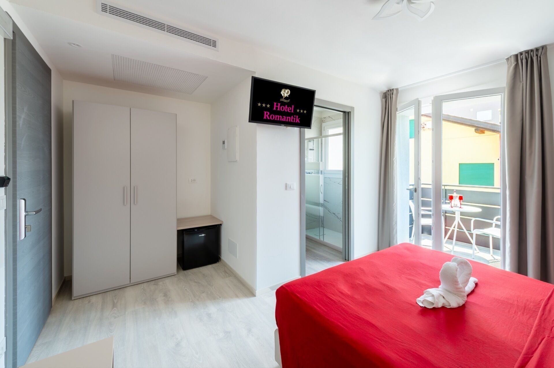 A hotel room with a red bed, a small desk, a wardrobe, a mini-fridge, a shower area, and a balcony with outdoor seating.