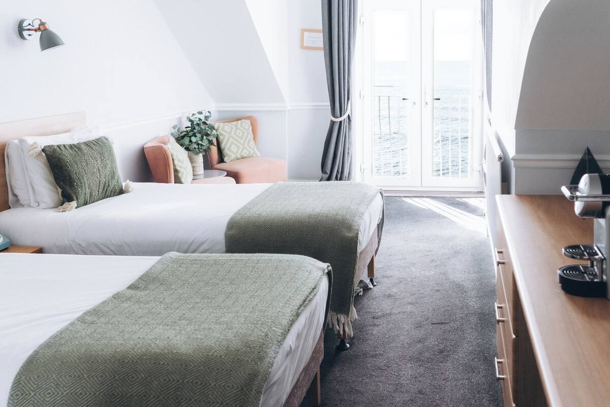 A hotel room with two beds, a wooden desk, a potted plant, and a view of the sea through a large window.