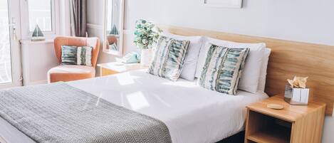 Habitación de hotel ordenada con una cama grande, una mesita de noche, una silla y una ventana con cortinas.