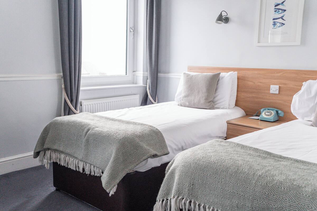 A hotel room with two beds, a wooden headboard, a bedside table with a telephone, and a window with curtains.
