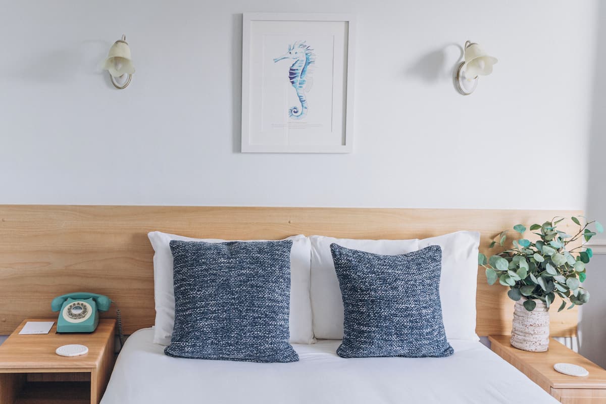 A neatly made bed with two pillows, a wooden headboard, a framed seahorse artwork, a vintage telephone, and a vase with green leaves on a bedside table.