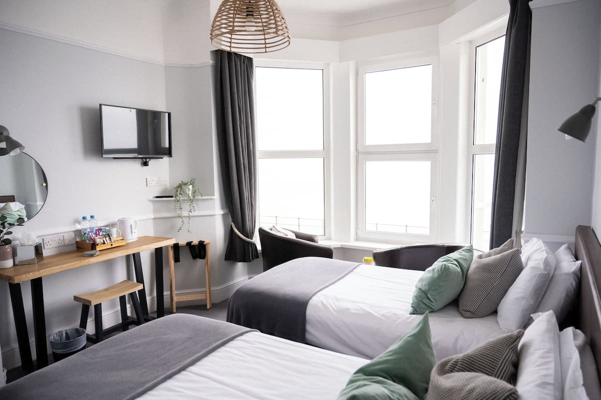 A modern hotel room with two beds, a desk, a TV, and large windows.