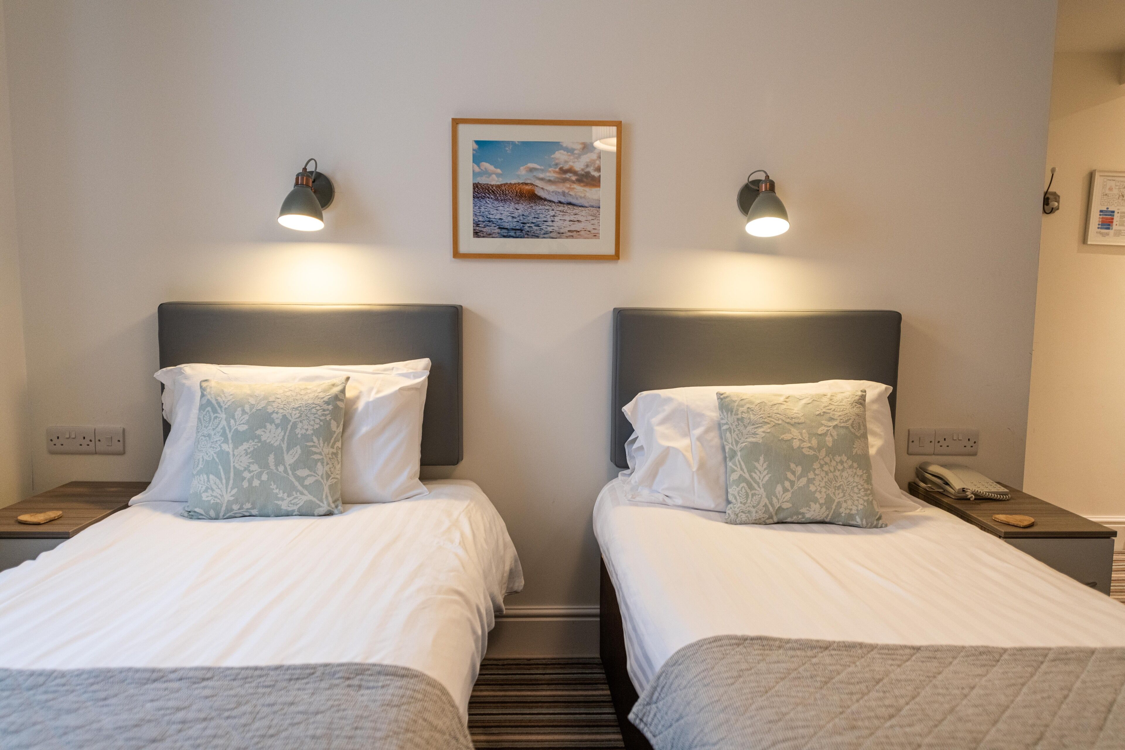 A hotel room with two single beds, each with a headboard, bedside tables, and a framed picture on the wall.