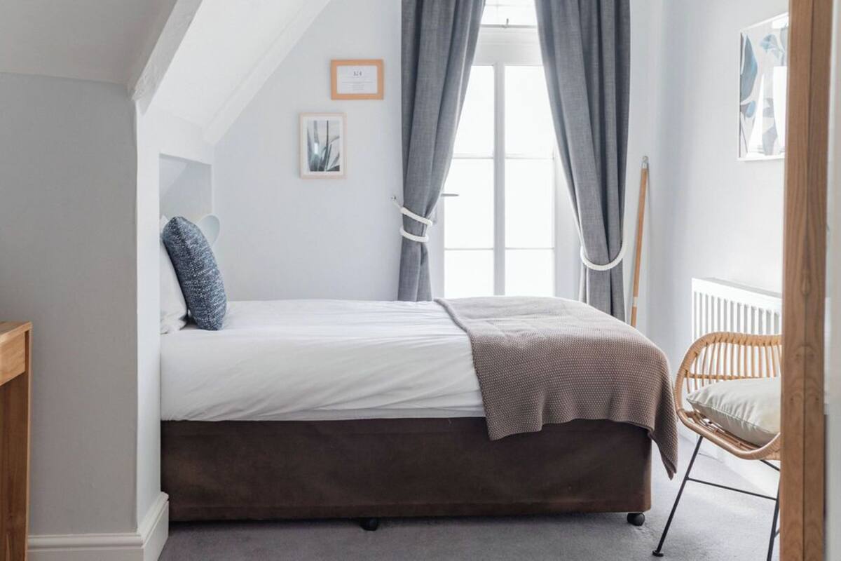 Een slaapkamer met een eenpersoonsbed, een raam met gordijnen, een houten stoel en een ingelijste foto aan de muur.