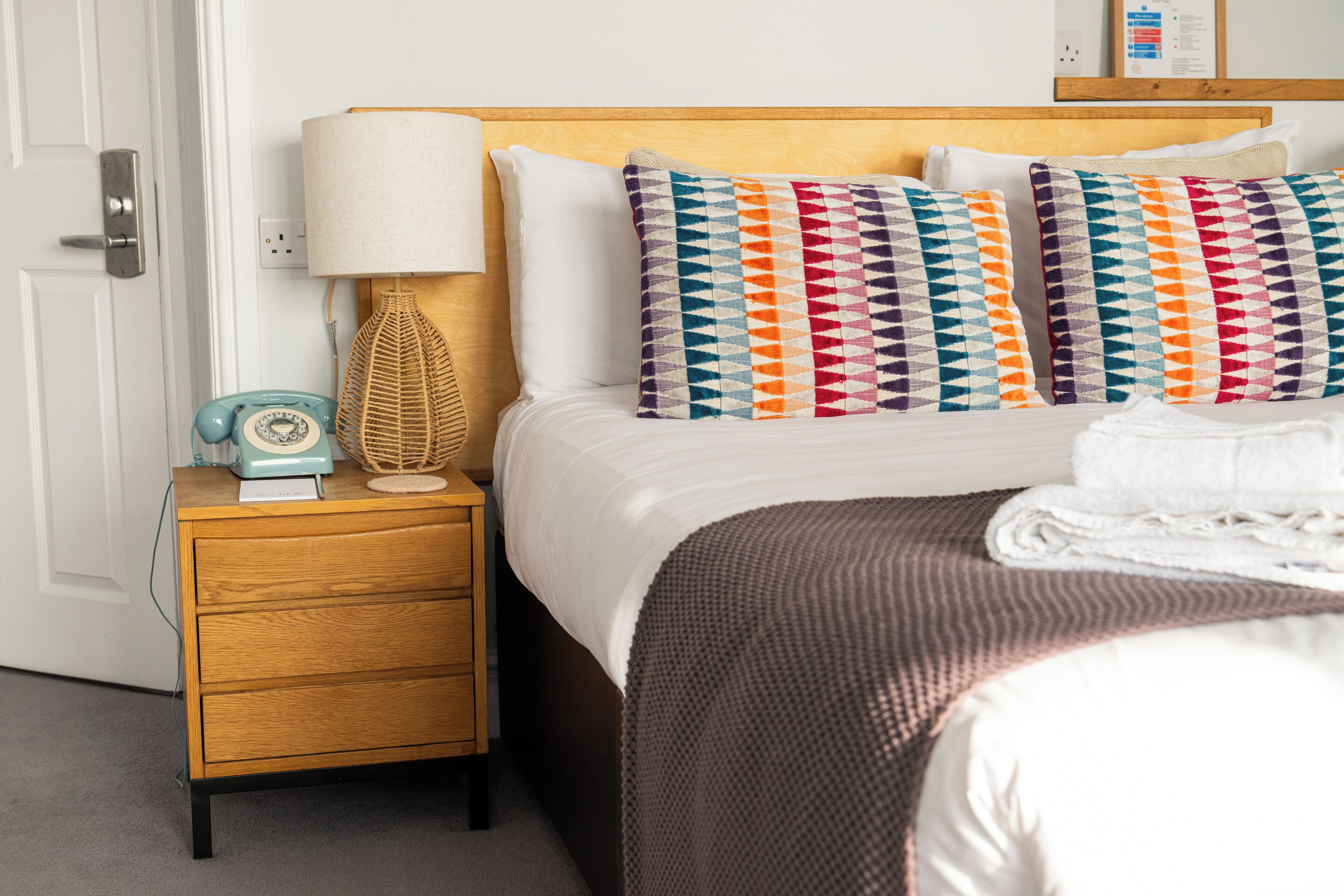 A neatly made bed with white linens, a wooden nightstand with a lamp and a vintage telephone, and a colorful geometric pillow.