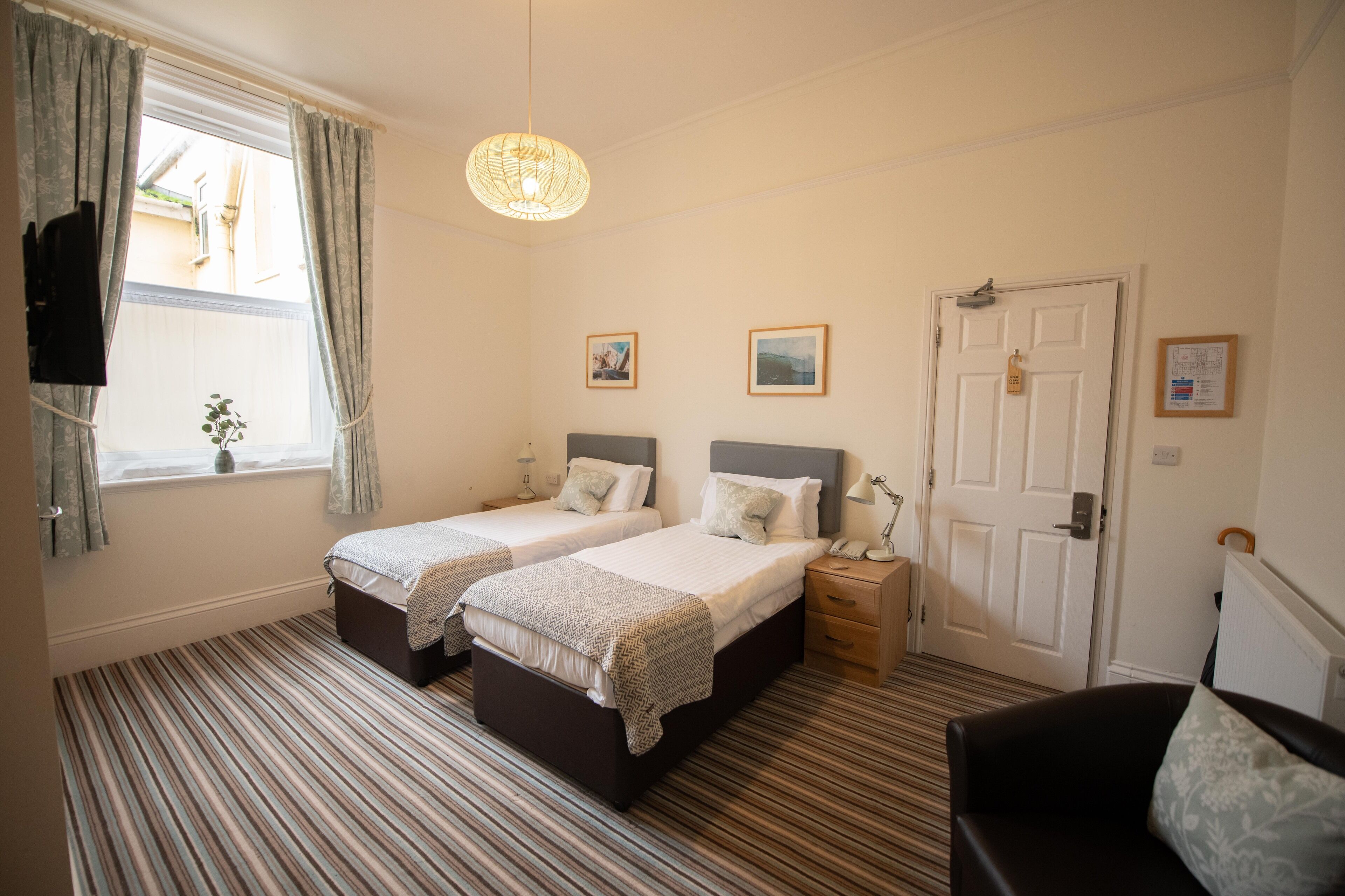 A hotel room with two beds, a large window with curtains, a hanging light fixture, and a door with a lock.