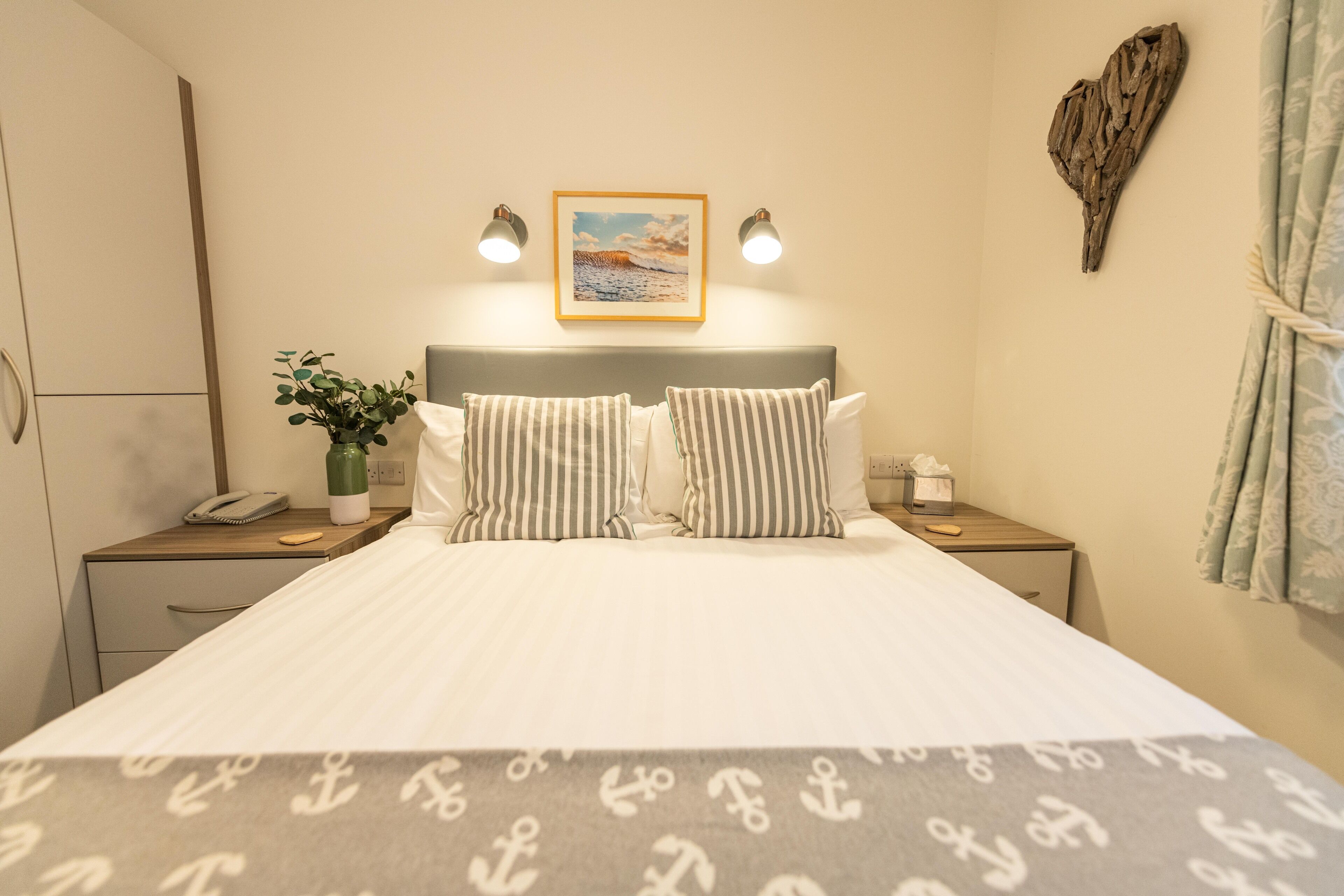 A neatly made bed with striped pillows, a bedside table with a phone and a bottle, a wall-mounted picture, and a decorative heart-shaped object.