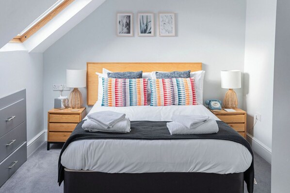 Un dormitorio moderno con techo inclinado, cabecera de madera y cama con almohadas de colores. Hay dos mesitas de noche con lámparas y un pequeño reloj.